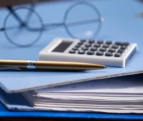 Black wire-framed spectacles, a white calculator and a gold pen lie on a light blue accounting ring-binder folder 