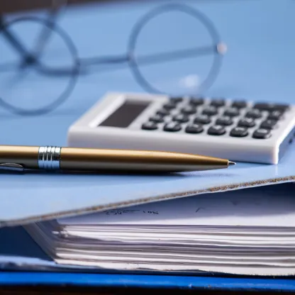 Black wire-framed spectacles, a white calculator and a gold pen lie on a light blue accounting ring-binder folder 